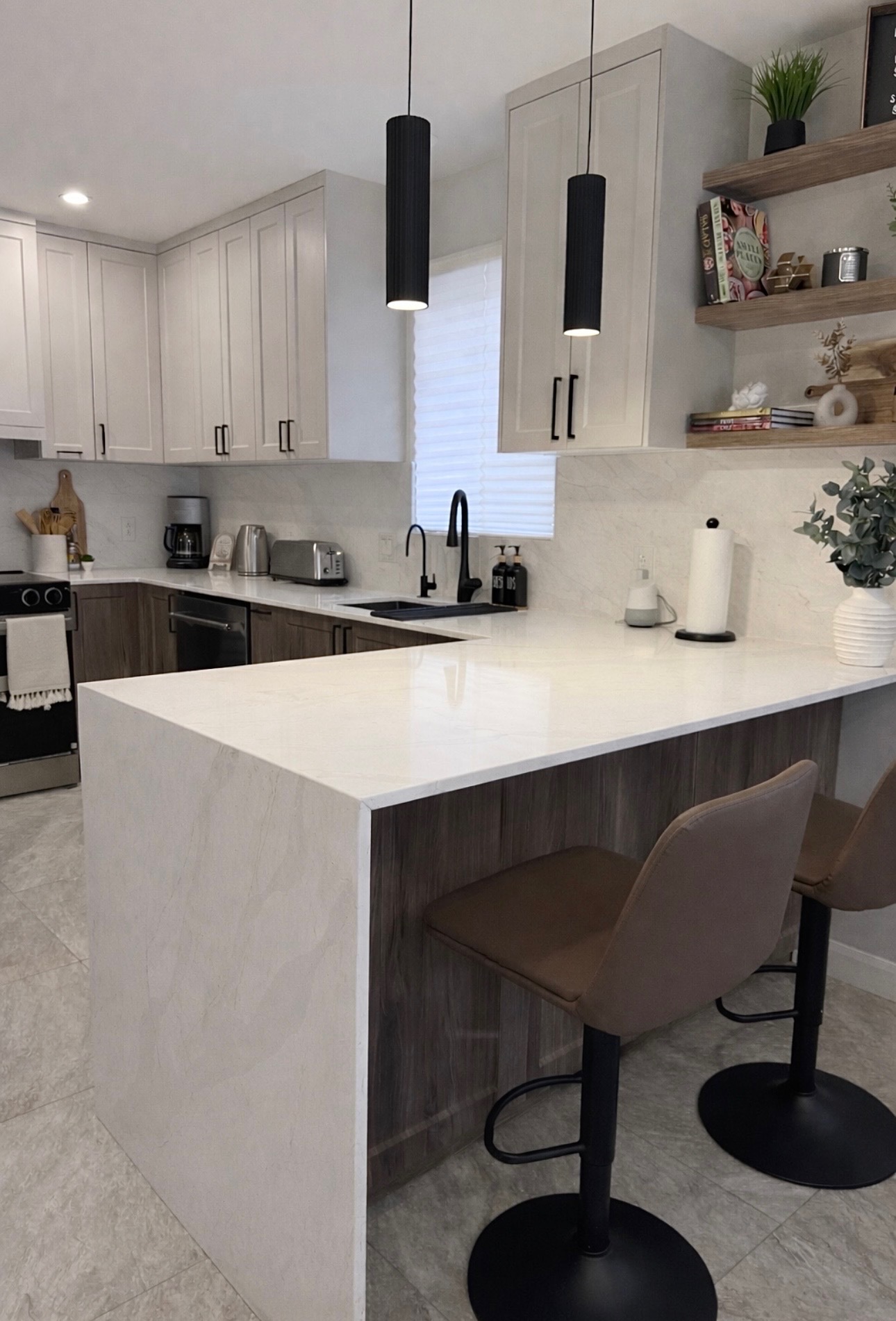 Cuisine ouverte Construction Altora avec péninsule en quartz blanc, armoires en noyer et façades hautes blanc cassé, suspensions cannelées noires.
