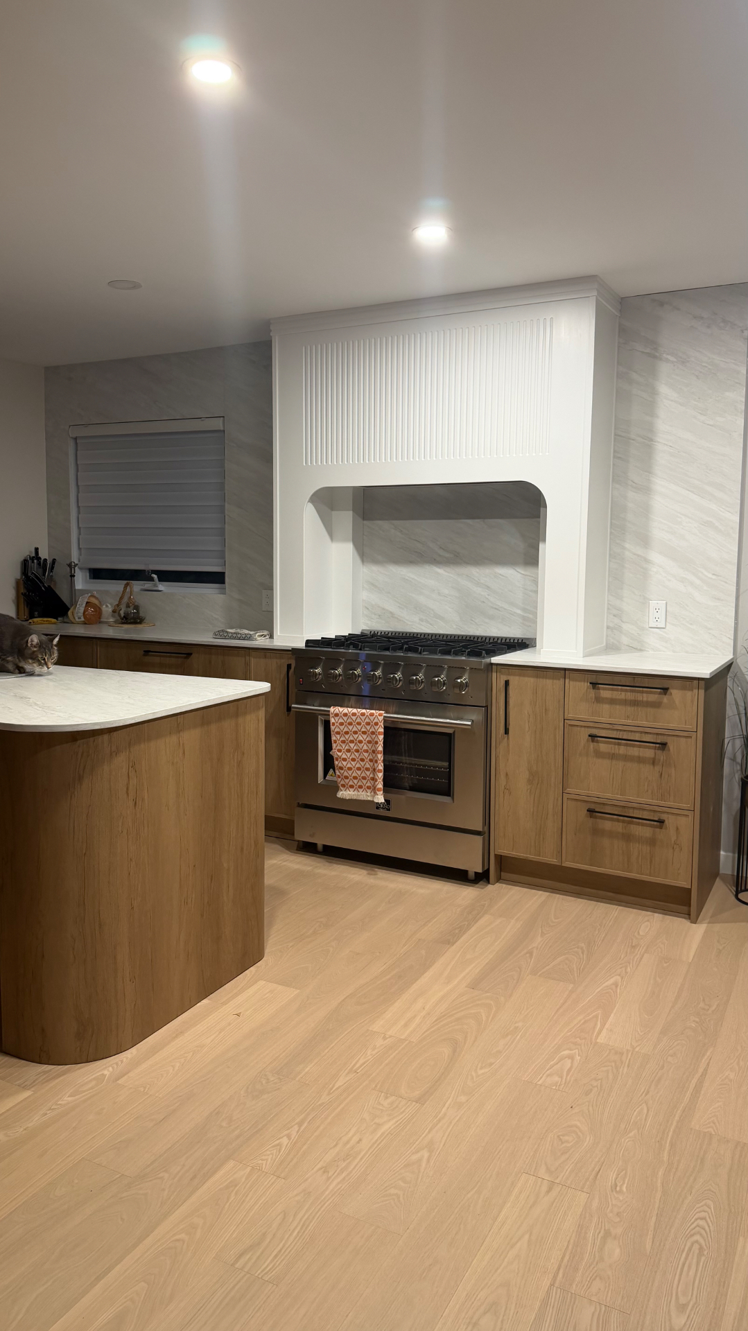 Vue de la cuisine Belmont depuis le salon — cuisinière encadrée par la hotte cannelée, îlot courbe en premier plan.