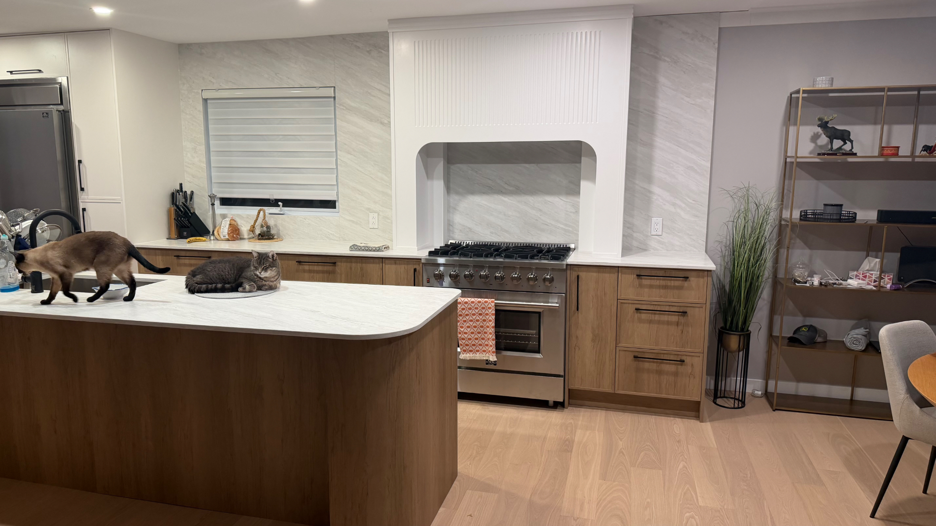 Vue élargie de la cuisine Belmont — îlot en chêne, comptoirs en marbre veiné, colonnes de rangement et étagère en laiton à droite.