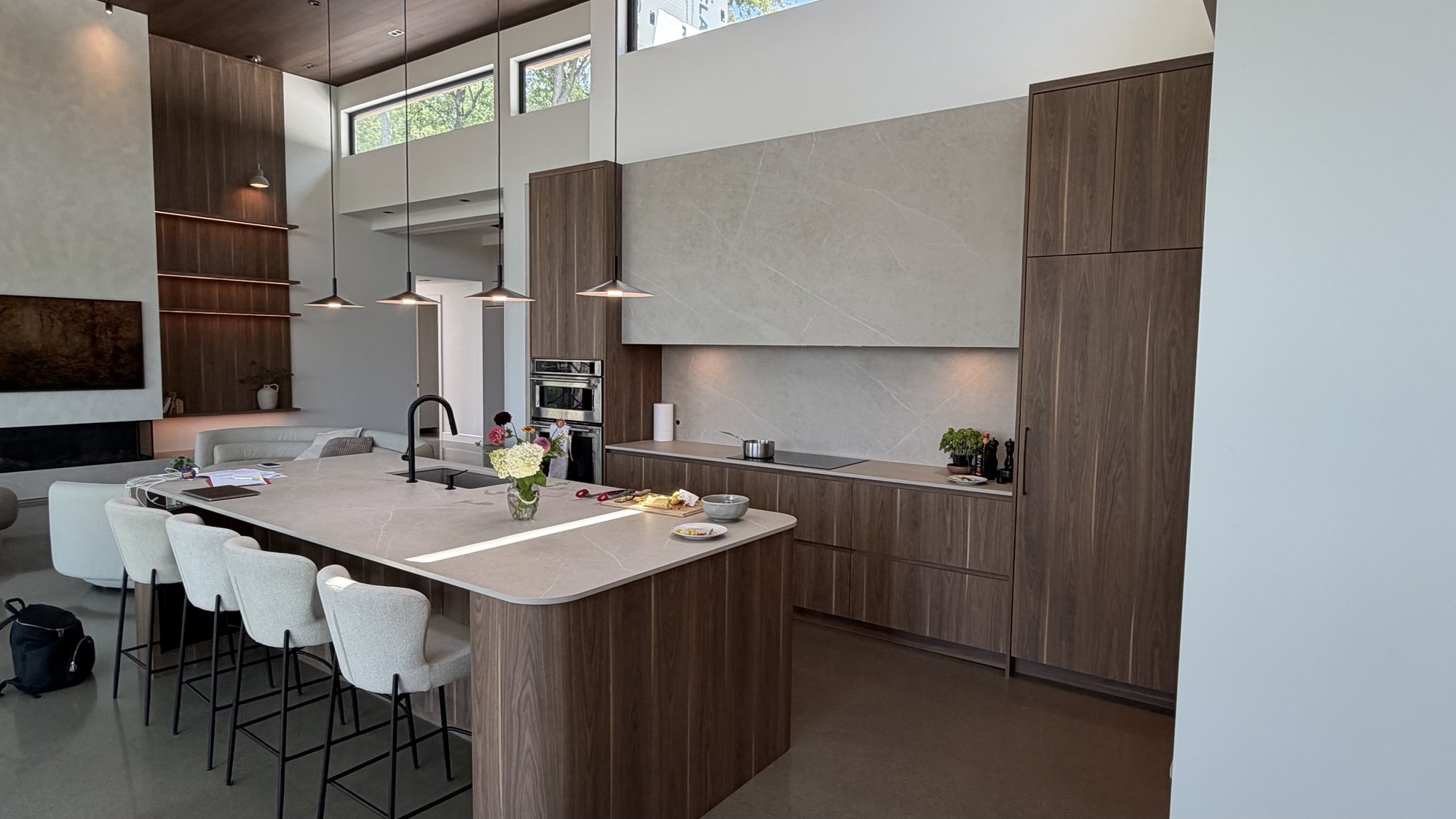 Cuisine à aire ouverte du Projet 30e Avenue à Laval — grand îlot en placage de noyer, dosseret et hotte pleine hauteur en Dekton, suspensions noires, plafond en bois massif à double hauteur.