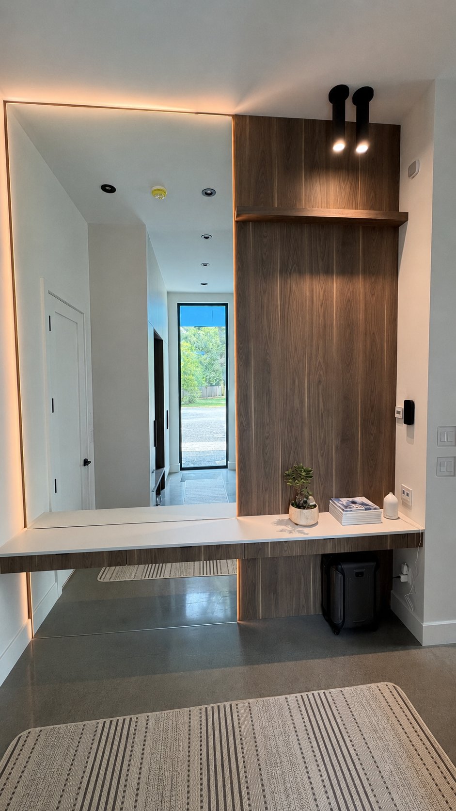 Entryway in the 30th Avenue Project — walnut-veneer console bench with a lit full-height mirror and a glazed door to the driveway.