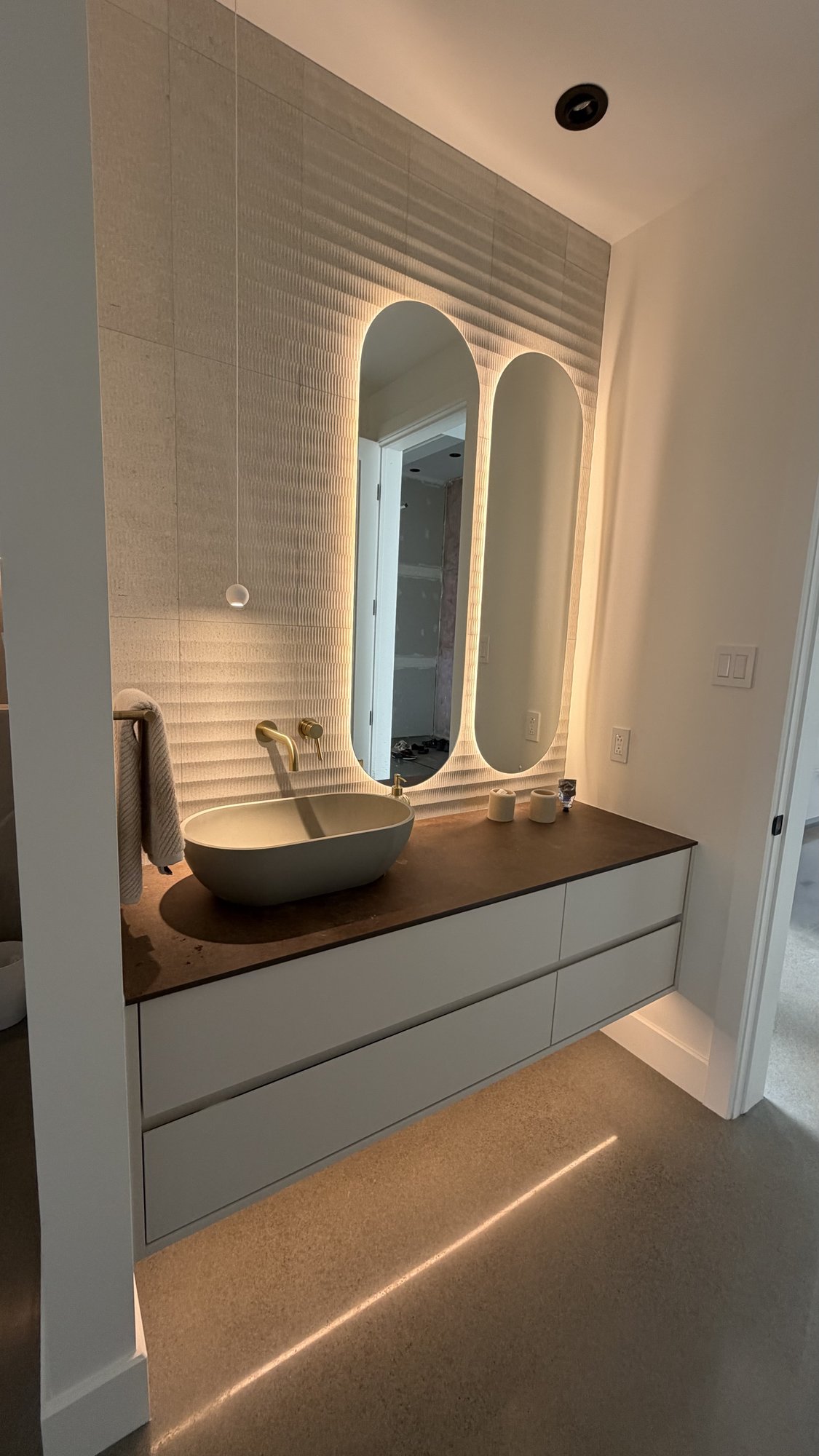 Powder room in the 30th Avenue Project — oval vessel sink on a Dekton counter, textured stone wall and a backlit double arched mirror.