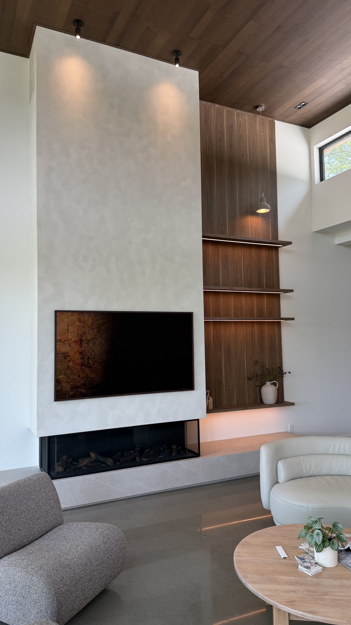 Living room in the 30th Avenue Project — three-sided fireplace finished in textured plaster, solid-wood ceiling rising to nearly 20 feet, walnut-veneer shelving with integrated lighting.
