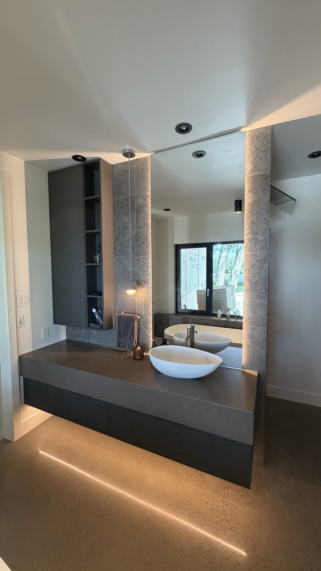 Primary suite double vanity, 30th Avenue Project — dark counter, textured-stone columns and a full-height backlit mirror.