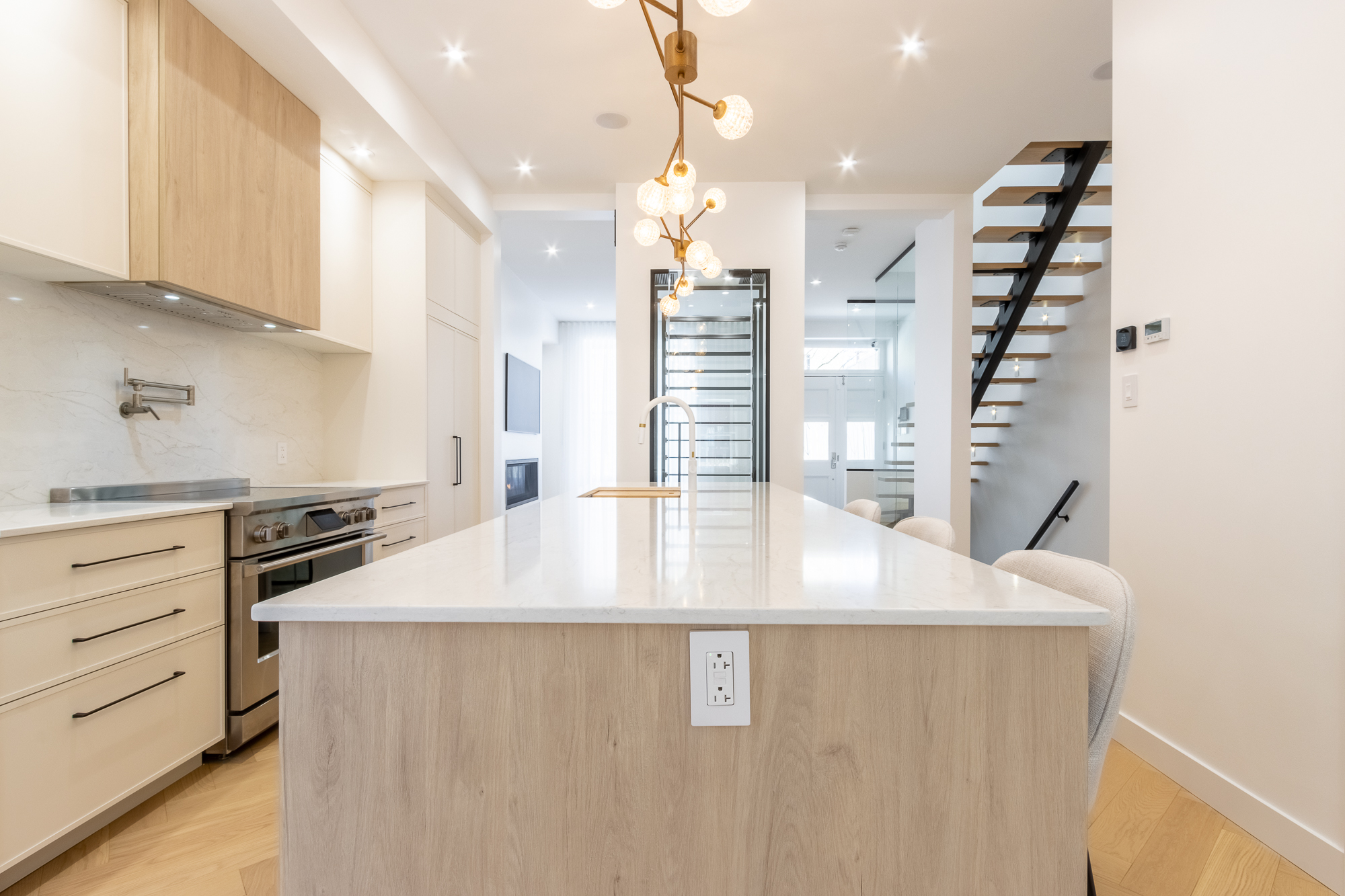 Cuisine Westmount avec long îlot à effet cascade en quartz veiné, lustre sputnik en laiton, cellier vitré et escalier flottant en arrière-plan.